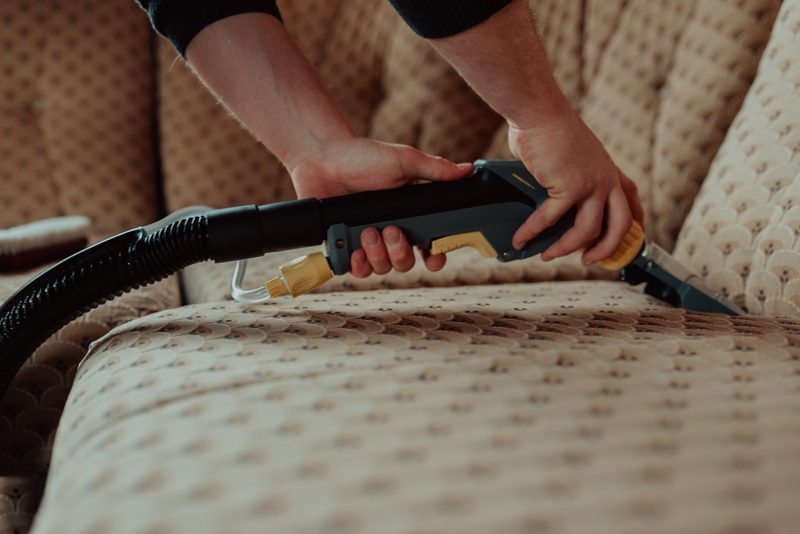 Close-up of housekeeper holding modern washing vacuum cleaner and cleaning dirty sofa with professionally detergent. Professional springclean at home concept